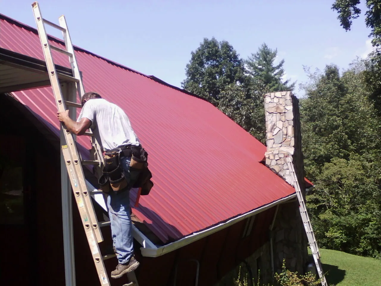 Roofer on a ladder beside a red metal roof and stone chimney — Soffit & Fascia Repair by Roofing Brothers in Apple River
